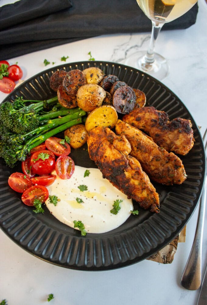 A black plate with grilled chicken, roasted potatoes, broccolini, cherry tomatoes, and a white sauce.