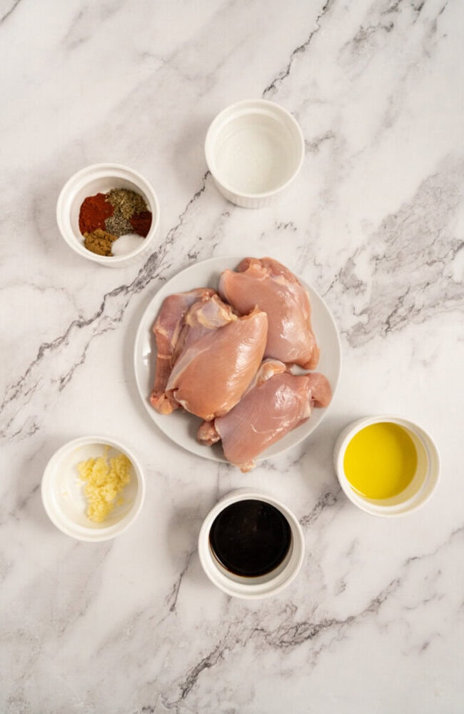 Raw chicken thighs on a plate, surrounded by bowls of oil, spices, vinegar, garlic, and soy sauce on a marble surface.