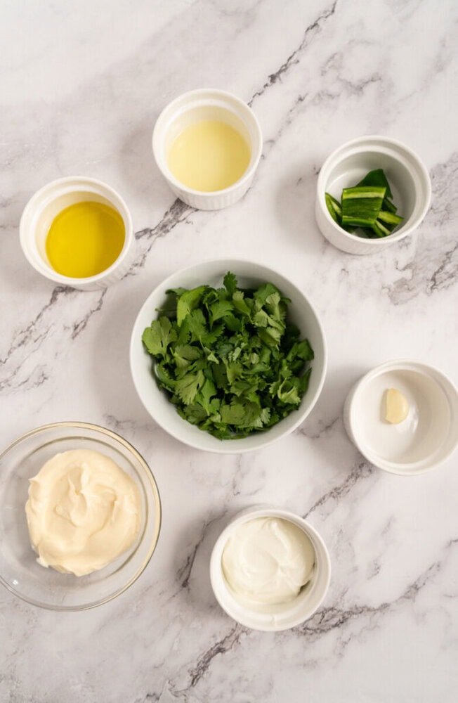 Bowls with mayonnaise, sour cream, cilantro, oil, lime, and a garlic clove on a marble surface.