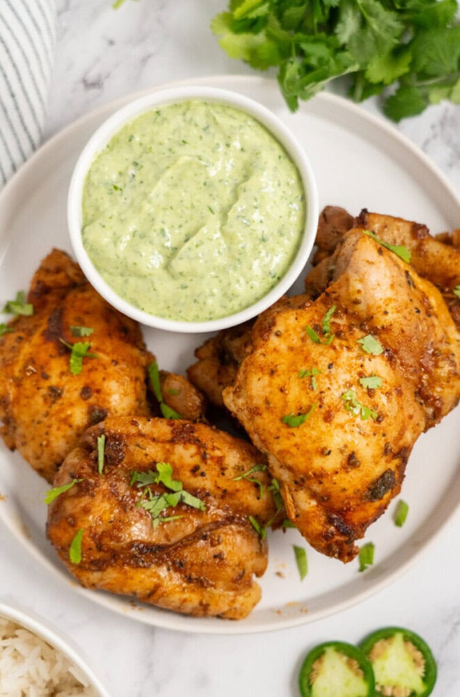 Three seasoned grilled chicken thighs garnished with herbs, served with a bowl of creamy green sauce on a plate.