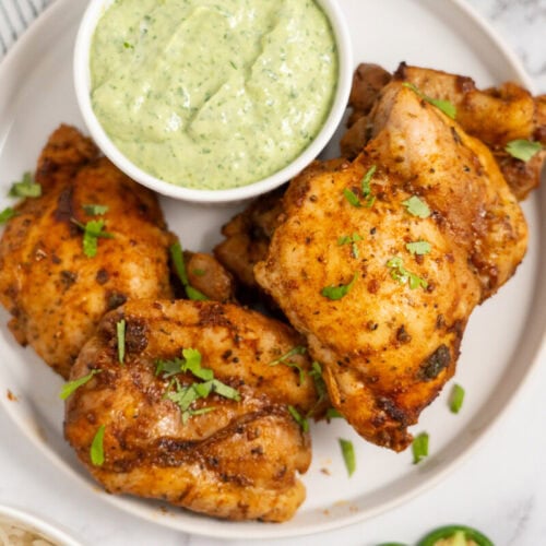 Plate of seasoned, cooked chicken thighs garnished with herbs, served with a bowl of creamy green sauce.
