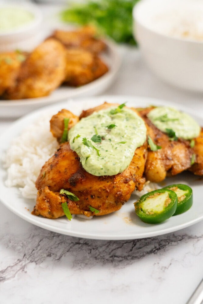 Seasoned chicken breast with creamy green sauce, served with white rice and sliced jalapeños on a white plate.