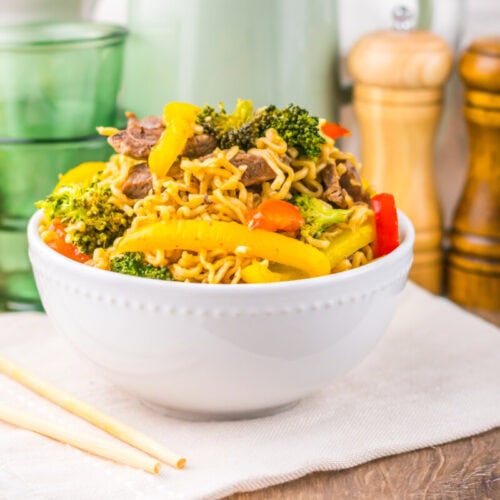A white bowl of stir-fried noodles with beef, broccoli, and colorful bell peppers beside chopsticks.