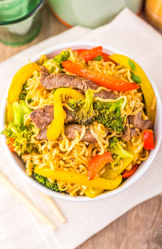 Bowl of beef stir-fry noodles with broccoli, red and yellow bell peppers, on a napkin with chopsticks.