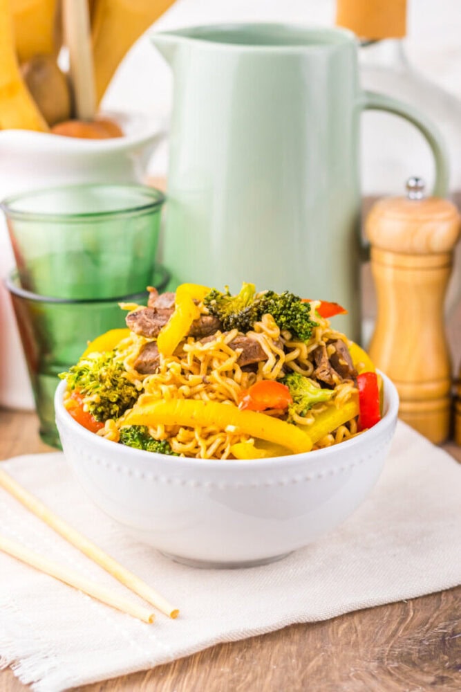 A white bowl of noodle stir-fry with beef, broccoli, and bell peppers on a table with chopsticks.