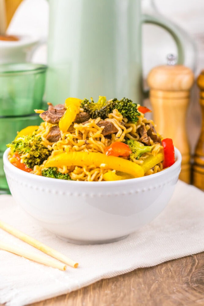 Bowl of stir-fried noodles with beef, broccoli, and colorful bell peppers on a table with chopsticks.