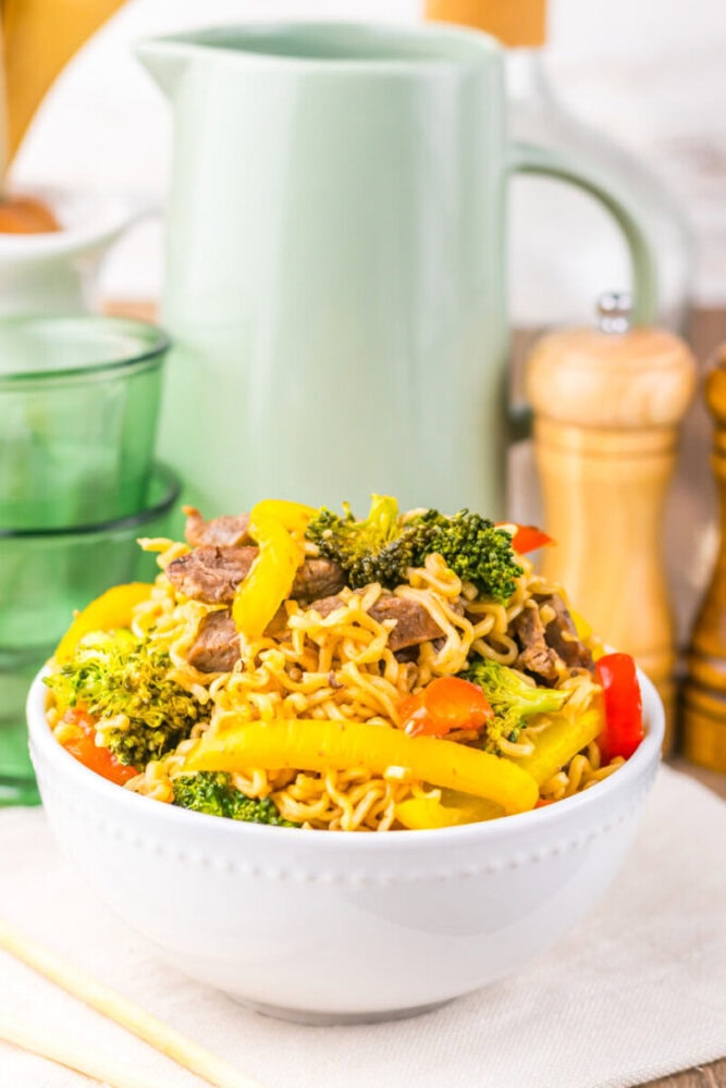 A bowl of stir-fried noodles with beef, broccoli, and colorful bell peppers on a table with kitchenware in the background.