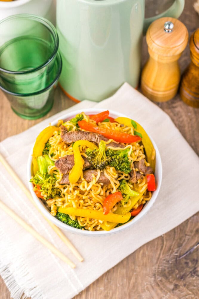 A bowl of stir-fried noodles with beef, broccoli, and bell peppers, next to chopsticks and green glasses.