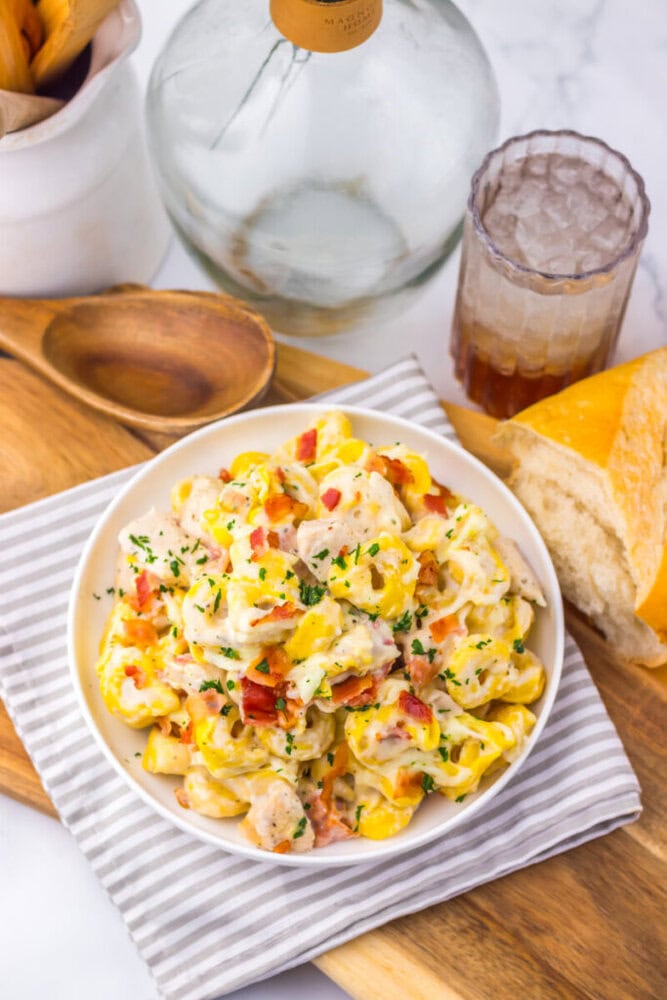 A bowl of creamy tortellini pasta with herbs, served with bread and iced drink on a wooden tray.