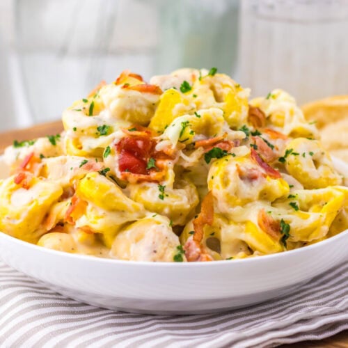 A bowl of creamy tortellini pasta with bacon bits and parsley, served with bread on the side.
