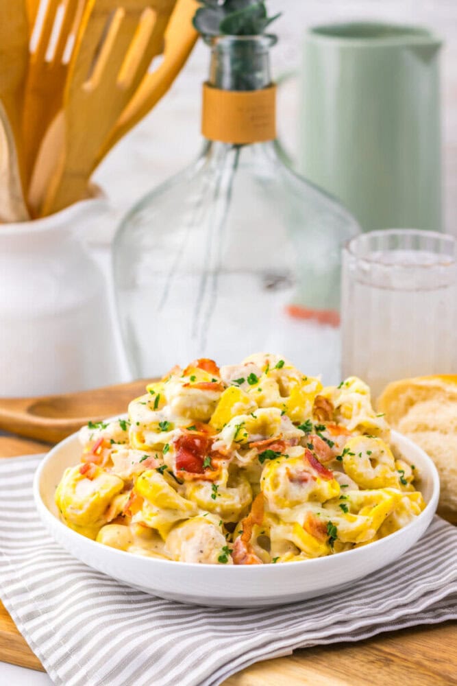 A bowl of creamy pasta with herbs and bacon sits on a striped cloth, beside bread and a glass of water.