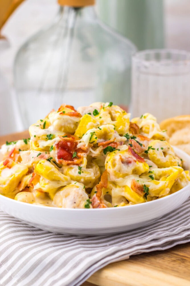 A bowl of creamy tortellini pasta with bacon bits and parsley, served on a striped napkin.