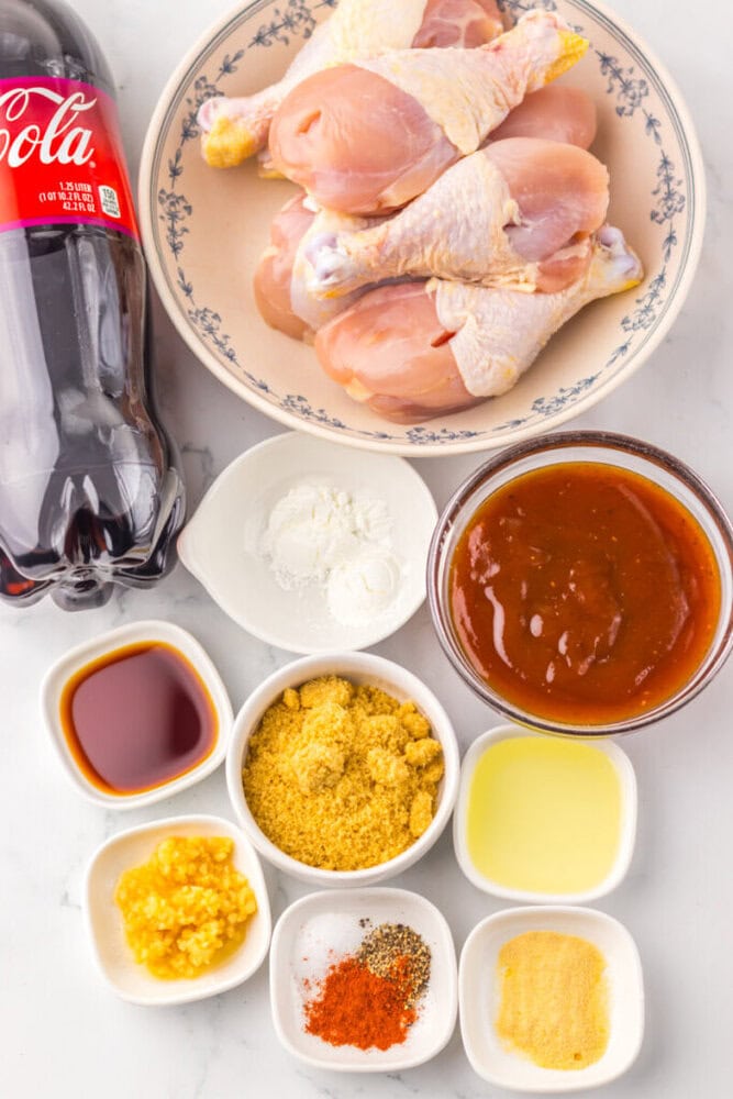 Raw chicken drumsticks on a plate with cola, sauces, seasonings, and spices in small bowls on a white surface.