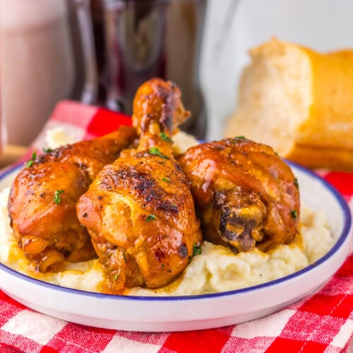 Three glazed chicken drumsticks on mashed potatoes, served on a plate with bread and a drink on the side.