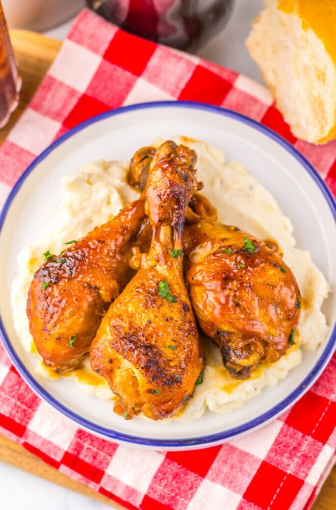 Three glazed chicken drumsticks served on creamy mashed potatoes in a white bowl on a red checkered cloth.