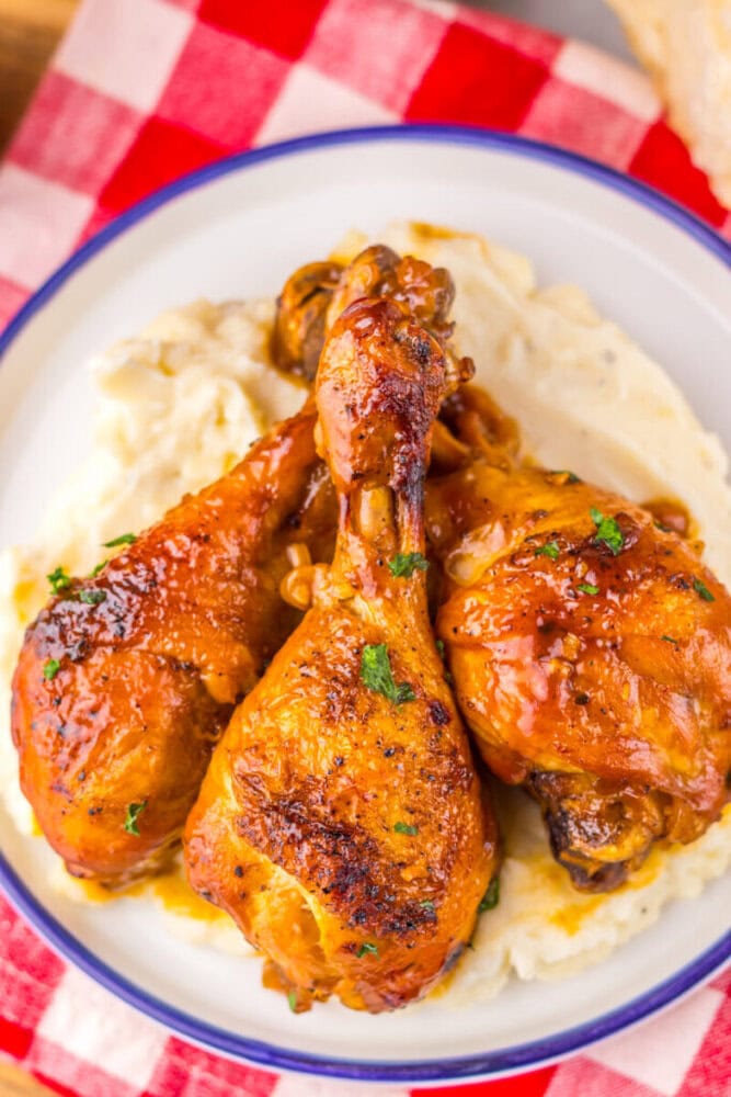 Three glazed chicken drumsticks on mashed potatoes, served on a white plate with a red checkered napkin.