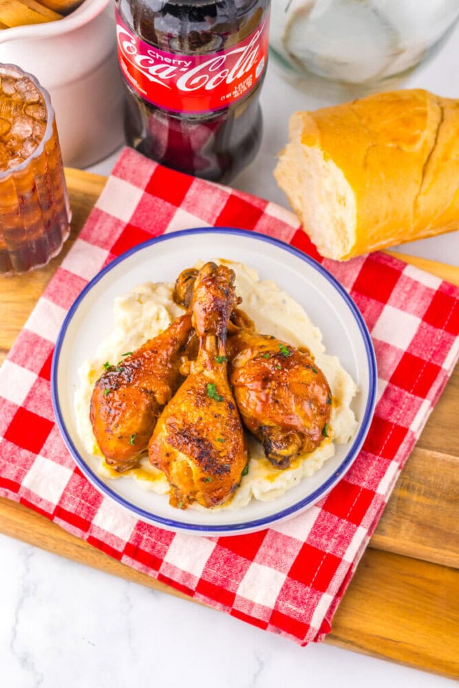 Three glazed chicken drumsticks on mashed potatoes, served with bread and a bottle of Cherry Coke on a red checkered cloth.