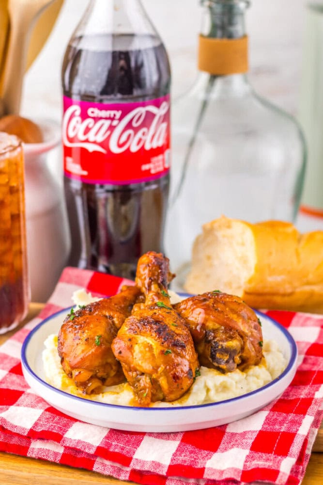 Three glazed chicken drumsticks on mashed potatoes, with bread and a bottle of Cherry Coca-Cola in the background.