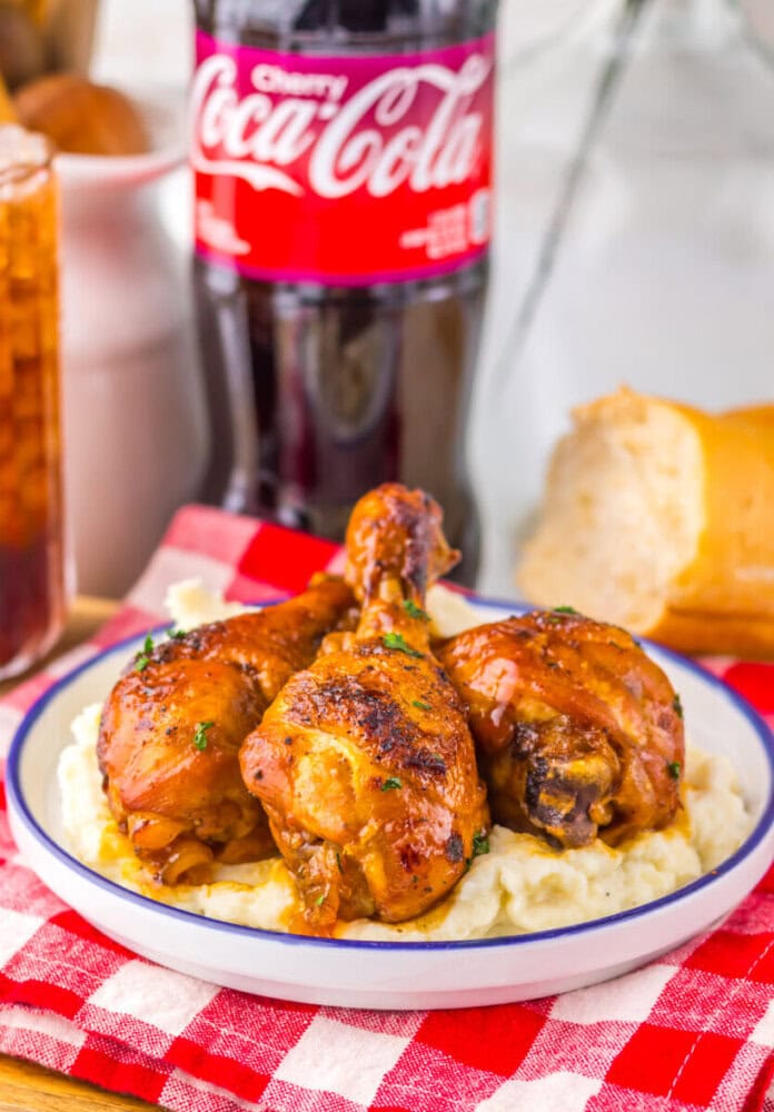 Three glazed chicken drumsticks on mashed potatoes, with a Coca-Cola bottle and bread in the background.