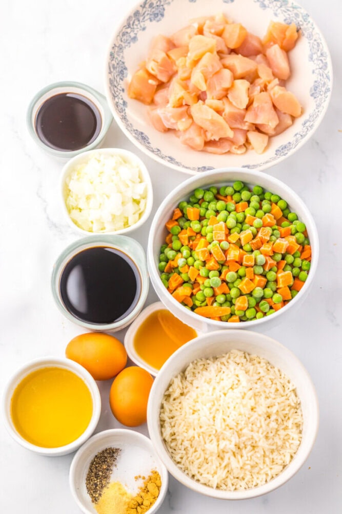 Bowls with chopped chicken, mixed vegetables, rice, eggs, seasonings, diced onion, and sauces on a white surface.