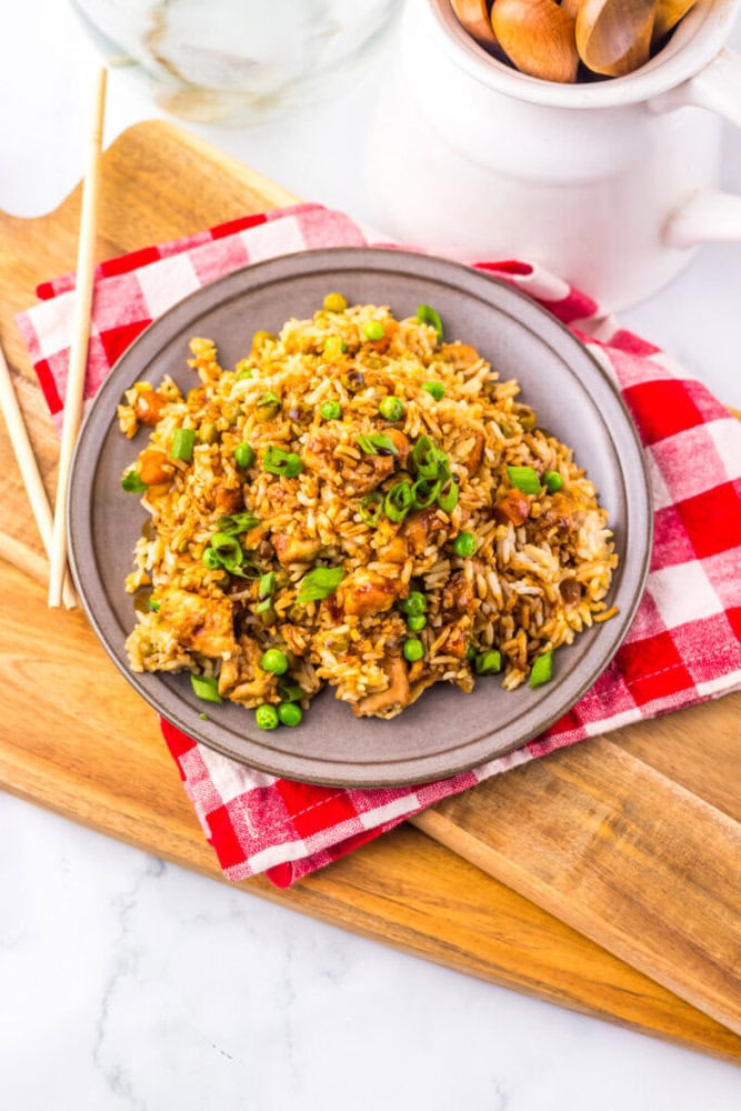 A plate of fried rice with vegetables on a gray plate, set on a red checkered cloth with chopsticks nearby.