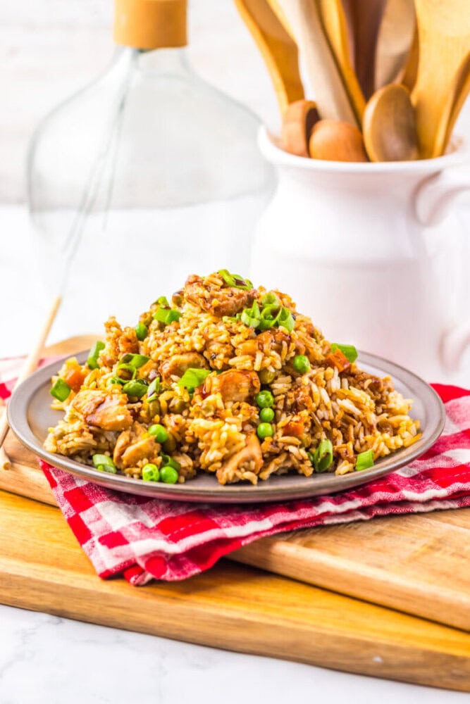 A plate of fried rice with chicken, peas, and carrots on a red napkin, kitchen utensils in the background.
