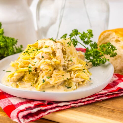 Creamy chicken and noodles garnished with parsley on a white plate, with bread and greens in the background.