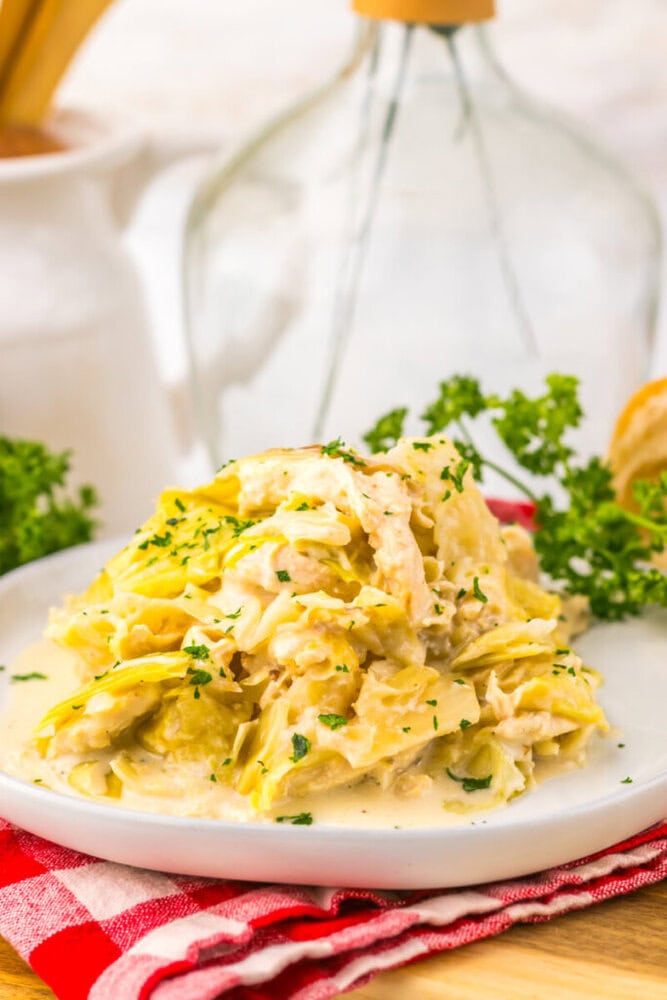 Creamy chicken and noodles garnished with parsley, served on a white plate with bread and herbs in the background.