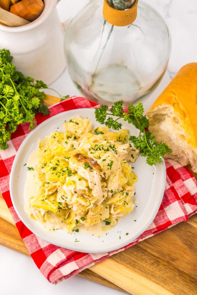 A plate of creamy pasta with chicken and herbs on a red checkered napkin, next to bread and a glass bottle.