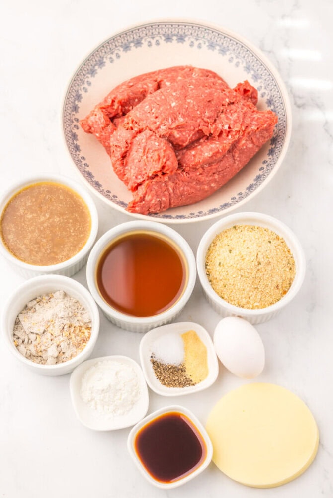 Ground beef, an egg, seasonings, sauces, breadcrumbs, and powders arranged in bowls on a white surface.