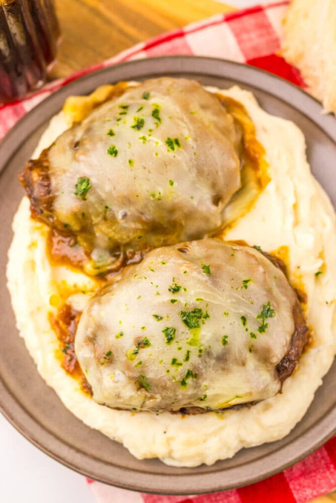 Two Salisbury steaks topped with melted cheese and herbs, served on mashed potatoes on a gray plate.