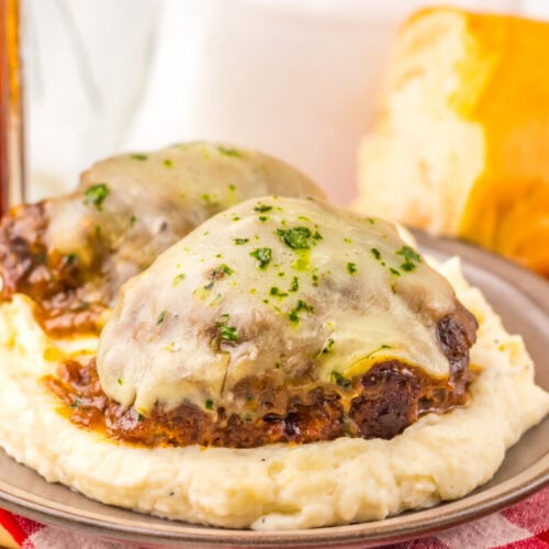 Two Salisbury steaks with melted cheese on mashed potatoes, served with bread and a drink in the background.