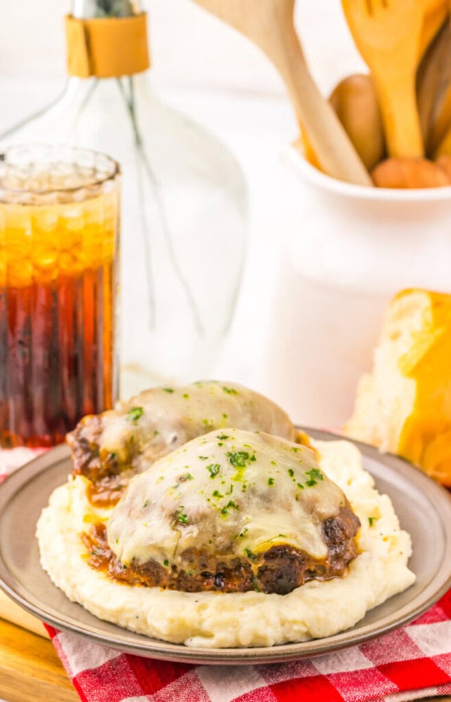 Two hamburger steaks with melted cheese on mashed potatoes, served with iced drink, bread, and utensils nearby.