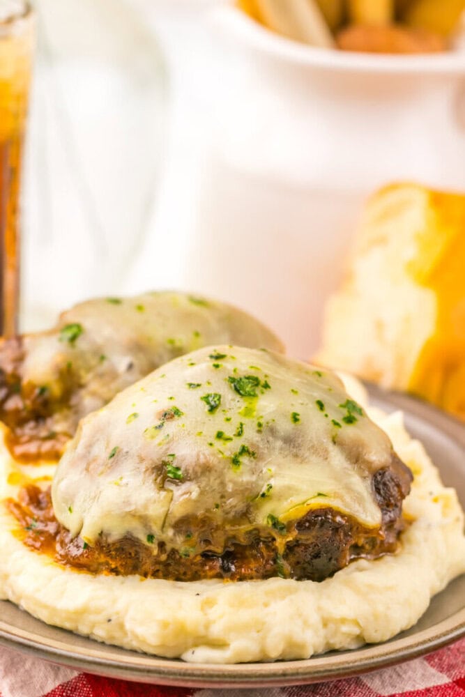 Two cheesy Salisbury steaks topped with herbs, served on a bed of creamy mashed potatoes on a plate.