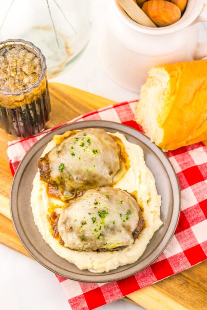 Two hamburger steaks with melted cheese on mashed potatoes, served with bread and a drink on a checkered napkin.