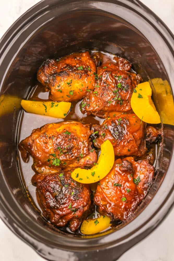 Slow cooker with glazed chicken thighs garnished with herbs and peach slices in sauce.