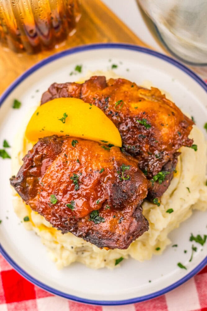Barbecued chicken thighs and a peach slice on mashed potatoes, garnished with herbs on a white plate.