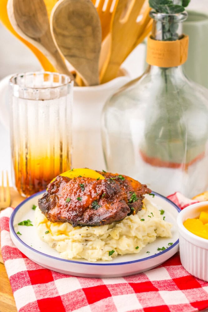 A plate of mashed potatoes topped with beef and peaches, next to a glass, bottle, and wooden utensils on a red checkered cloth.