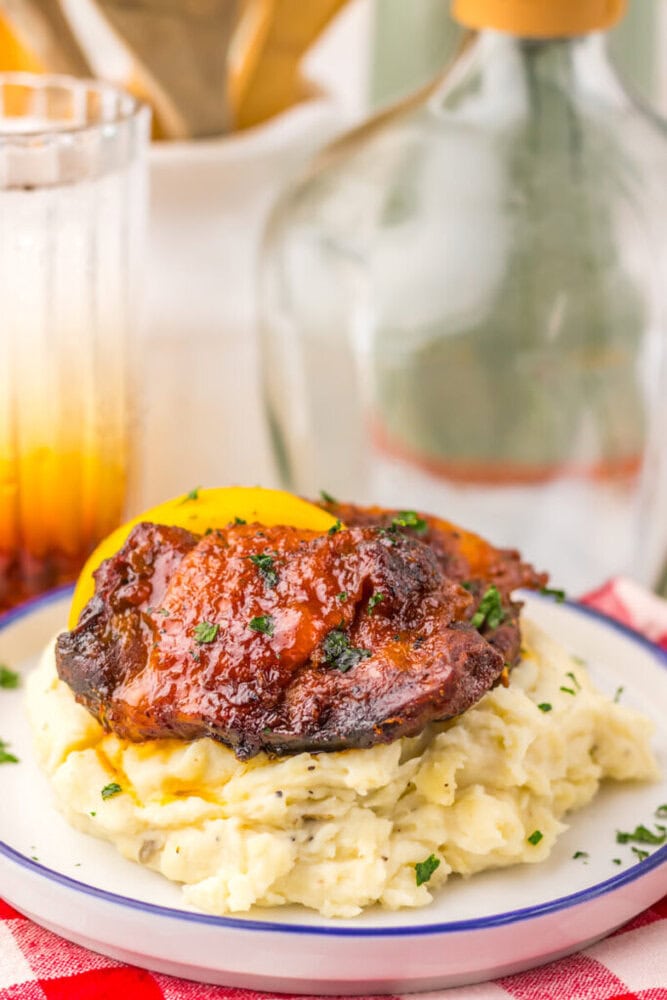 Barbecued chicken on mashed potatoes, garnished with herbs, served on a plate with a drink in the background.