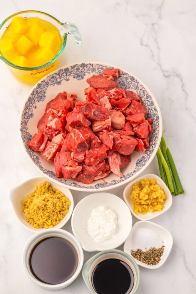 Raw diced beef, pineapple chunks, green onions, and various sauces and seasonings in small bowls on a marble surface.