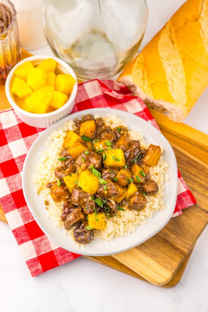A plate of rice topped with beef and pineapple chunks, next to a baguette and a bowl of cubed pineapple.