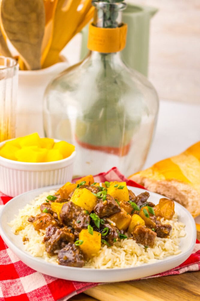 A plate of rice topped with beef stew and pineapple, with bread and a bowl of pineapple chunks beside it.