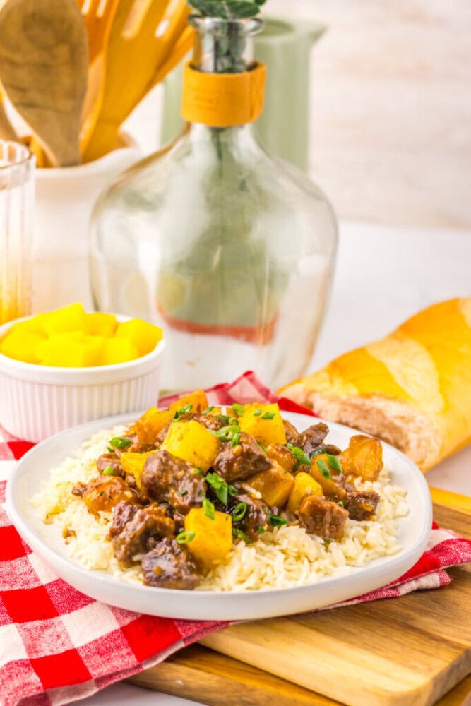Plate of beef stew with pineapple served over rice, next to bread and a bowl of pineapple on a red checkered cloth.