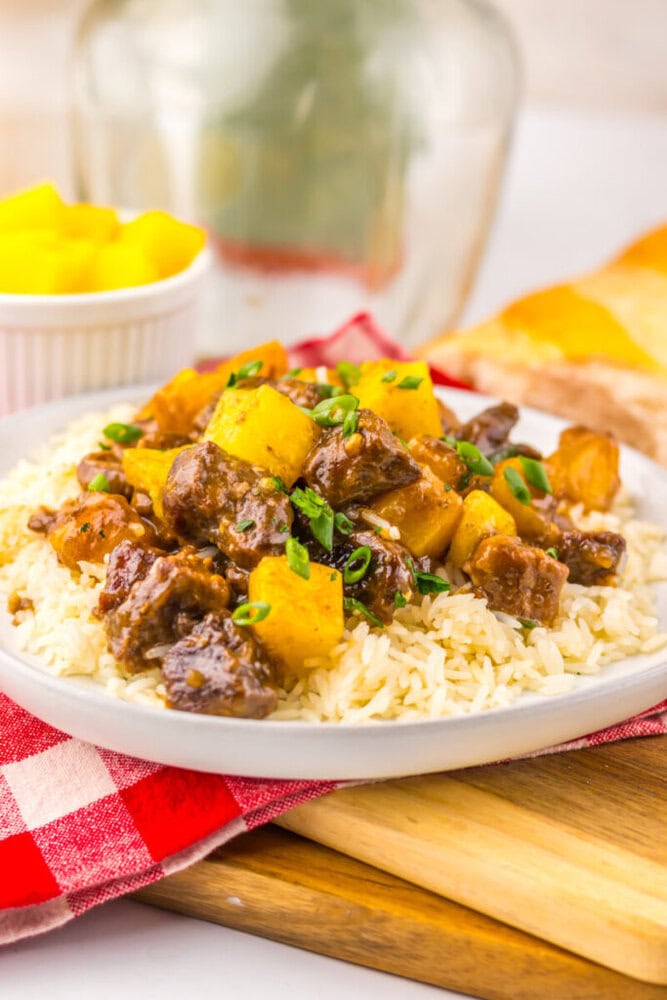 Beef stew with pineapple and green onions served over rice on a white plate, with mango cubes in the background.