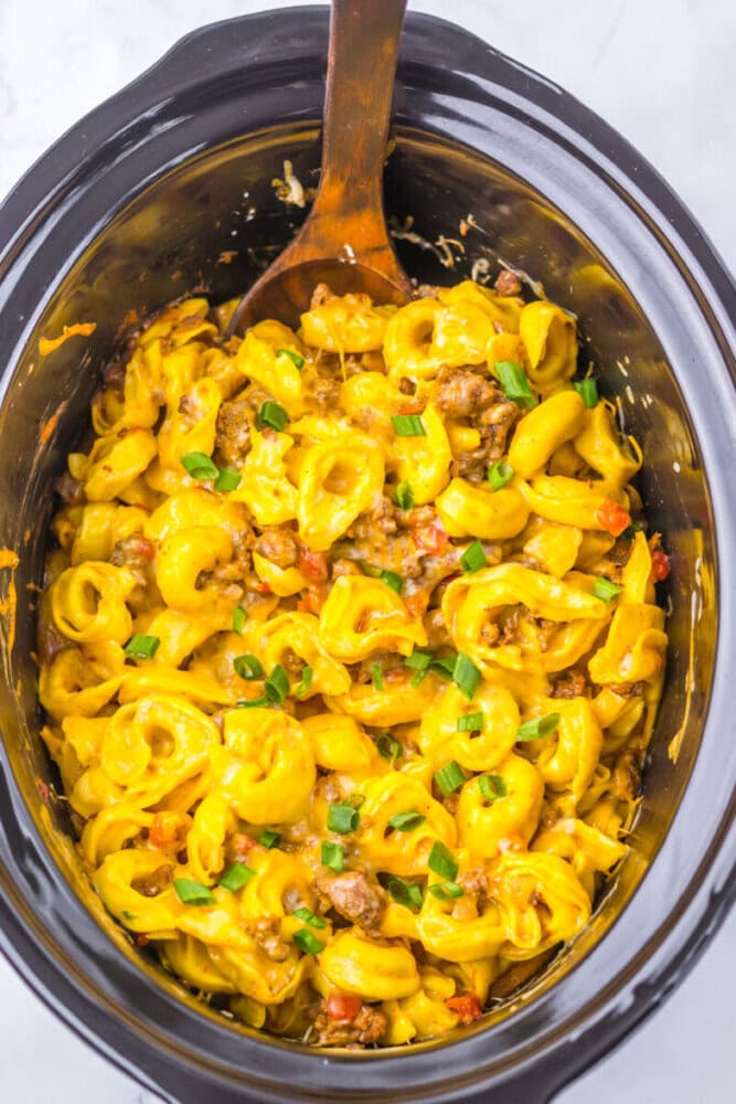 Cheesy tortellini with ground beef, bell peppers, and green onions in a slow cooker, stirred with a wooden spoon.