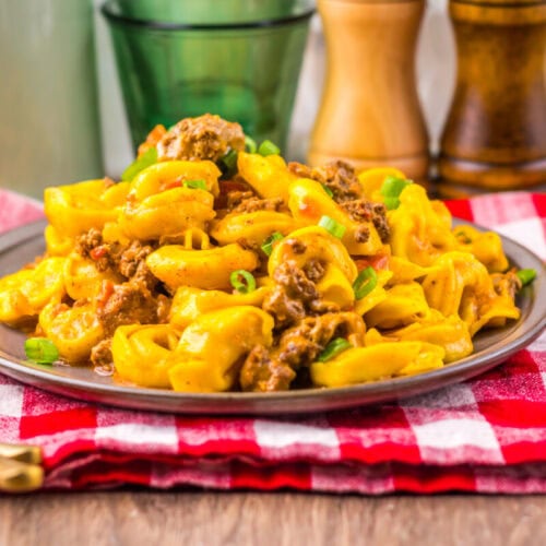 A plate of cheesy pasta with ground beef on a red checkered cloth, with condiments and a tomato nearby.