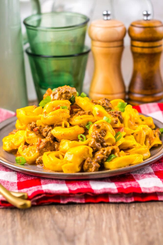 Plate of cheesy tortellini with ground meat and green onions on a table, with salt and pepper shakers in background.