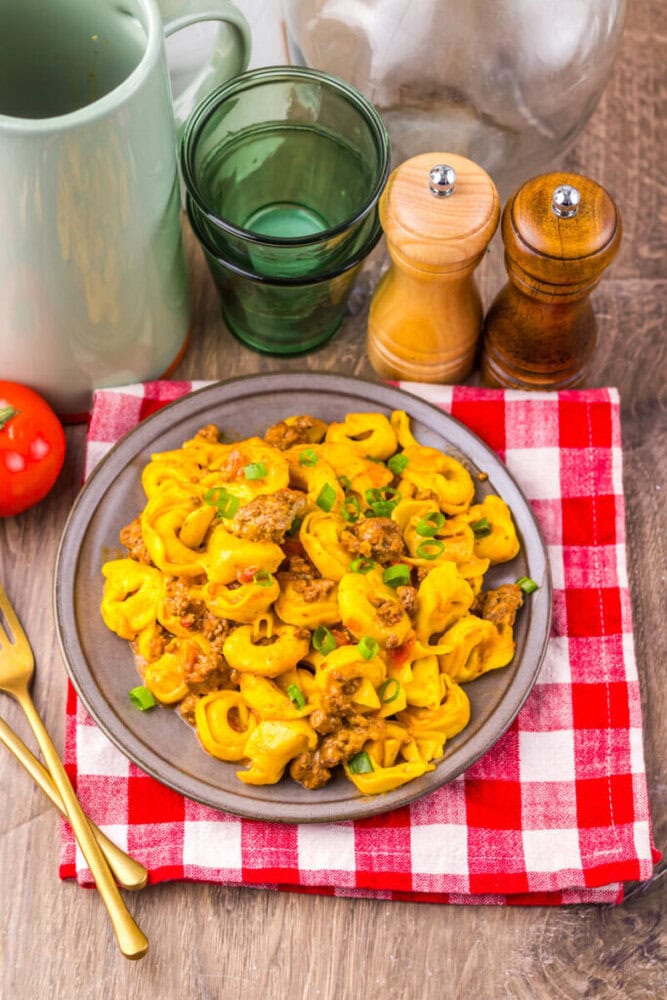 A plate of cheesy pasta with ground meat on a red checkered napkin, with utensils and glassware nearby.