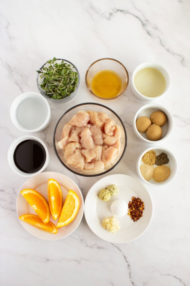 Top-down view of various bowls containing raw diced chicken, sauces, spices, orange slices, and microgreens.