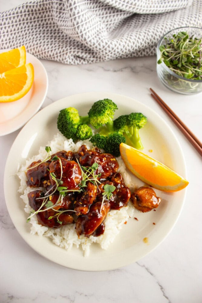 Plate of glazed chicken on rice with broccoli, microgreens, and an orange slice; chopsticks and orange on side.